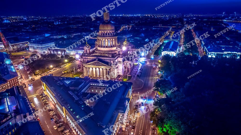 На фотографии запечатлён ночной вид на Исаакиевский собор в Санкт-Петербурге. Величественное здание собора подсвечено тёплым светом, подчёркивающим его архитектурную красоту. Вокруг собора расположены оживлённые улицы и исторические здания. Световые акценты создают атмосферу ночной жизни города.